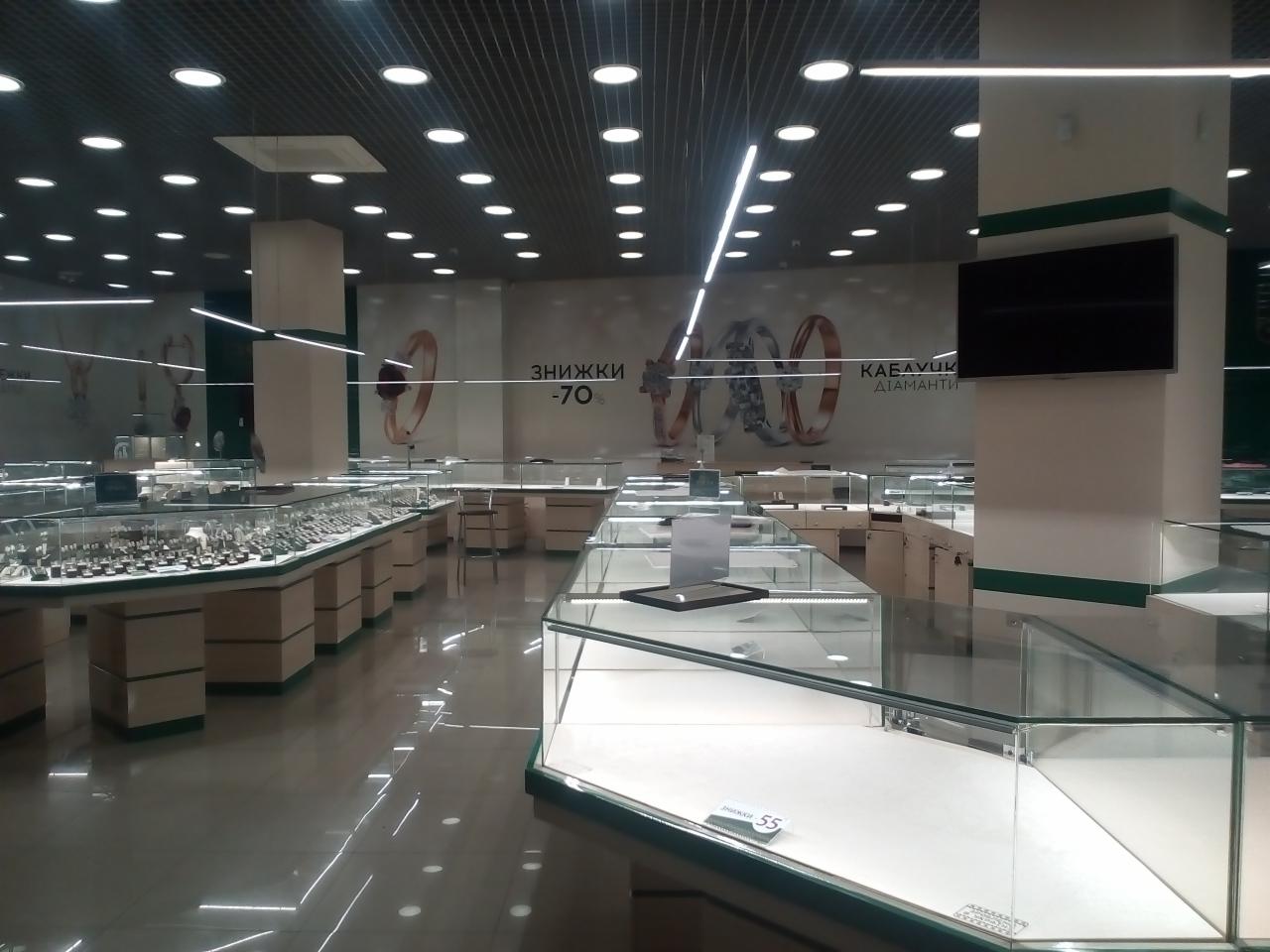 Wnętrze przestronnego sklepu jubilerskiego z licznymi szklanymi witrynami eksponującymi biżuterię, jasne oświetlenie sufitowe i plakat z promocją -70% w tle.
