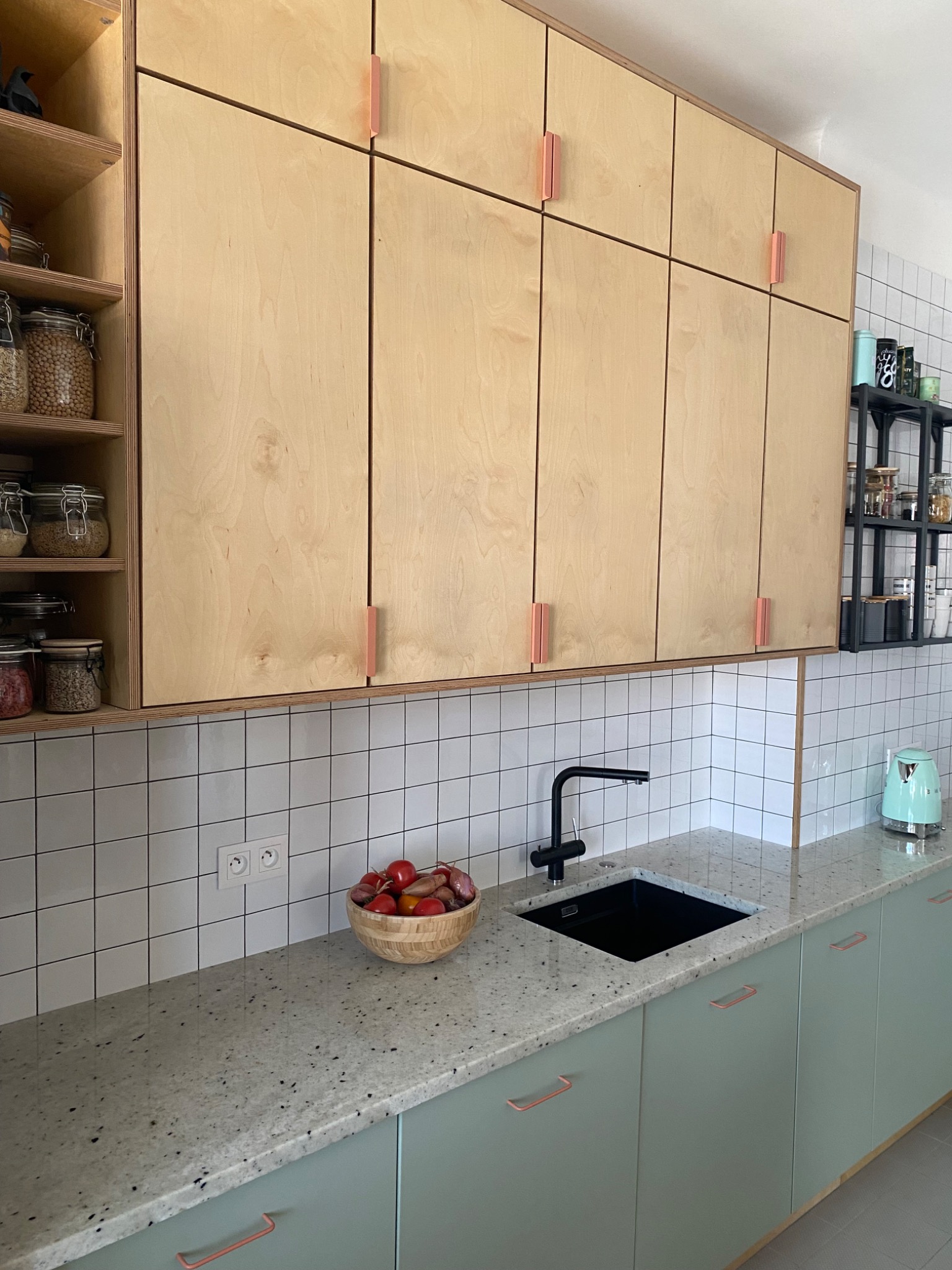 Nowoczesna kuchnia z jasnymi, drewnianymi szafkami górnymi z minimalistycznymi, miedzianymi uchwytami, zielonymi szafkami dolnymi i blatem z szarego lastryko, uzupełniona czarną armaturą i białymi...