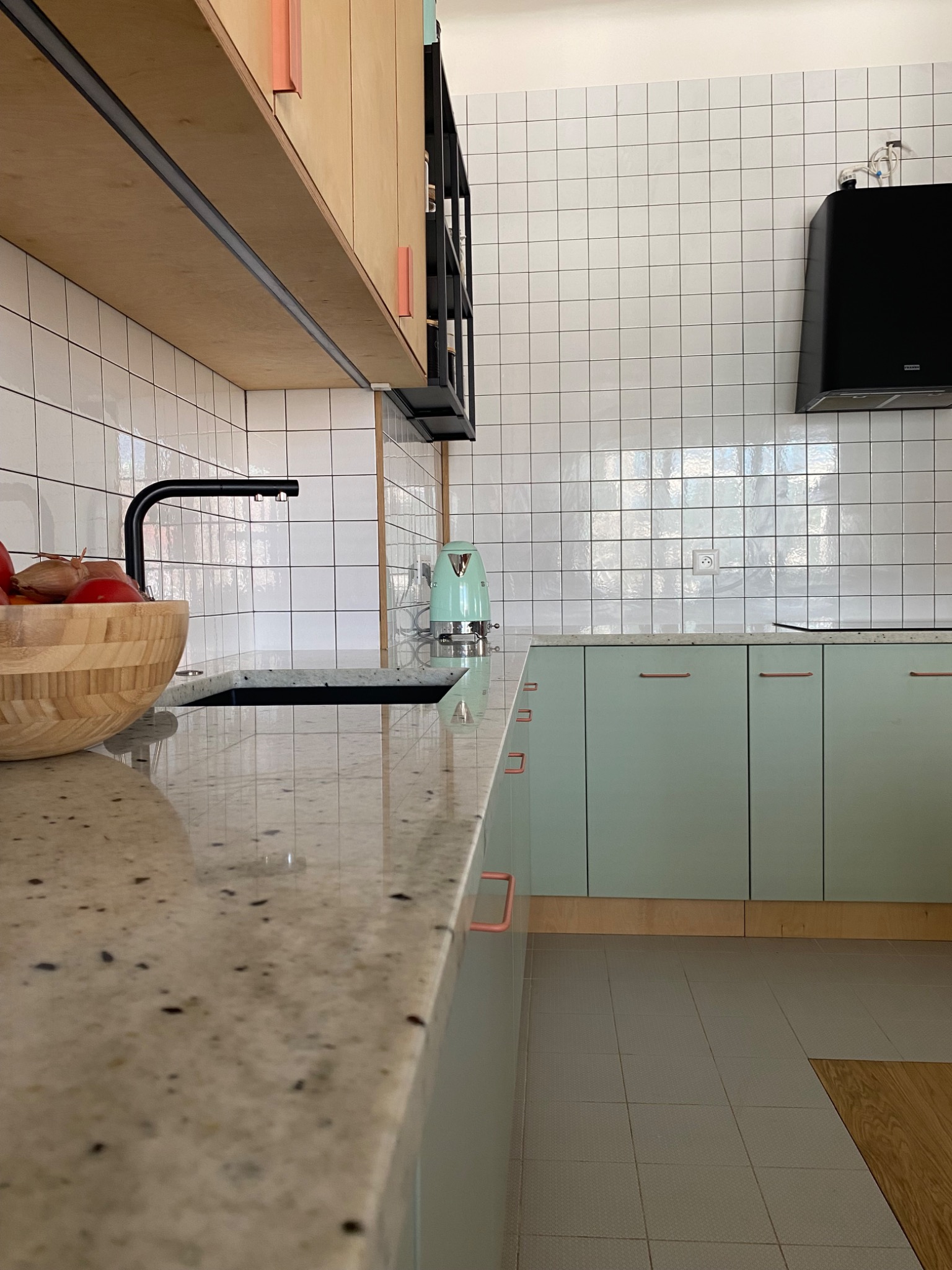 Fragment nowoczesnej kuchni z jasnymi, pistacjowymi szafkami z miedzianymi uchwytami, blatem z jasnego granitu, białymi kafelkami i czarną armaturą, uzupełniony drewnianą misą z owocami...