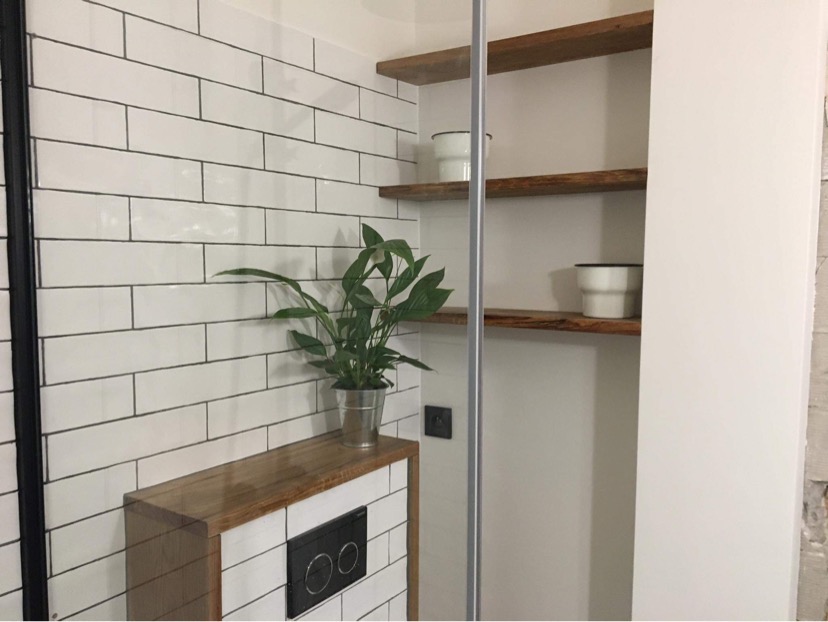 Nowoczesna łazienka z białymi płytkami, drewnianym blatem nad zabudowaną spłuczką i półkami z surowego drewna, na których stoją białe doniczki.