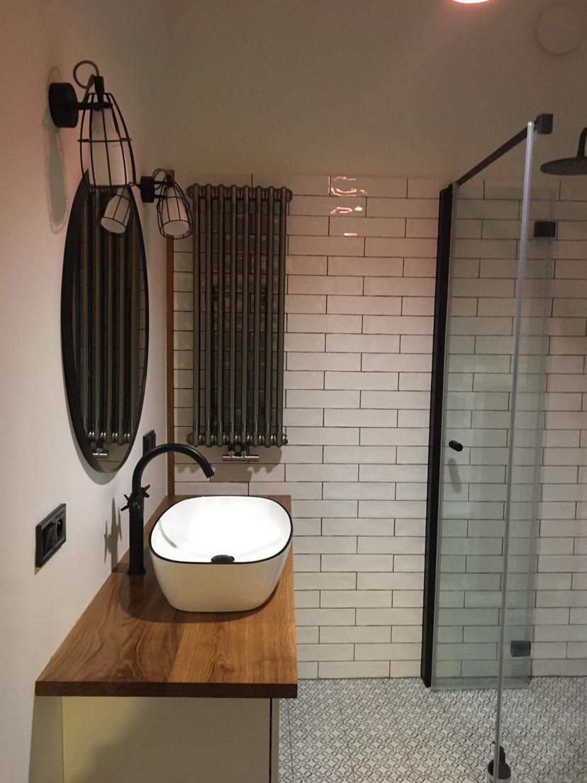Nowoczesna łazienka z owalnym lustrem w czarnej ramie, białą umywalką nablatową, drewnianym blatem i kabiną prysznicową z czarnymi profilami.