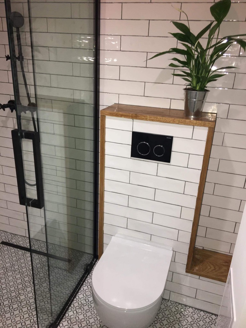 Nowoczesna łazienka z podwieszaną toaletą w obudowie z drewnianym blatem, nad którą stoi roślina w metalowej doniczce, kabina prysznicowa z czarnymi elementami, białe płytki i wzorzysta podłoga.