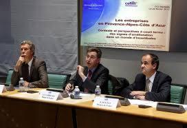 Trzej mężczyźni w garniturach siedzą za stołem podczas konferencji. Na stole widoczne mikrofony, butelki wody i plakietki z nazwiskami. W tle baner z napisem w języku francuskim.