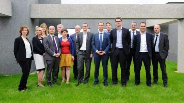 Grupa kilkunastu prawników, kobiety i mężczyźni, w strojach formalnych, stojąca na trawie przed nowoczesnym budynkiem z betonowymi elementami architektonicznymi.