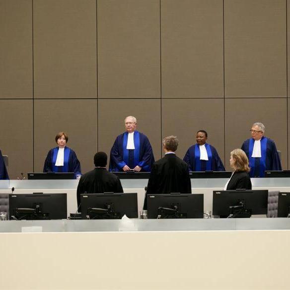 Sala sądowa z sędziami w togach podczas posiedzenia. Ustawione monitory przed każdym miejscem. Neutralne tło. Ujęcie z perspektywy publiczności.