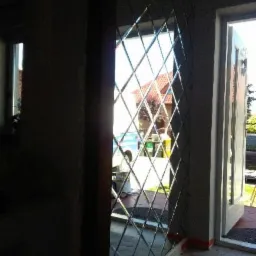 Ściana wyłożona dekoracyjnymi, romboidalnymi lustrami, odbijająca widok na zewnątrz z domem i pojazdami, widoczna częściowo otwarta biała rama drzwiowa.