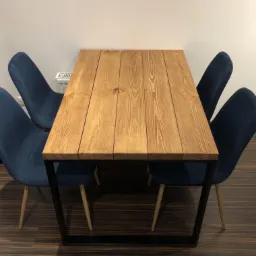 Stół z blatem z jasnego drewna klejonego z desek, na czarnych metalowych nogach, otoczony czterema krzesłami tapicerowanymi granatowym welurem na jasnych drewnianych nogach, na podłodze z paneli...