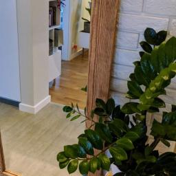 FaceWood - Duże lustro w drewnianej ramie z efektem postarzanego drewna, odbijające wnętrze mieszkania z białą biblioteczką i drewnianą podłogą, obok lustra roślina doniczkowa na okrągłym stoliku.