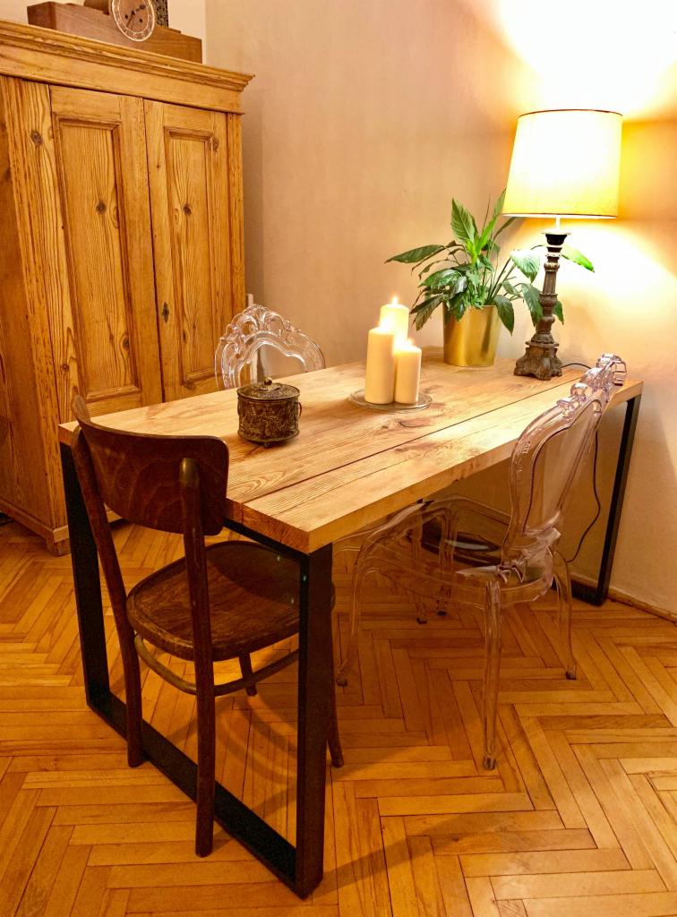 Stół z blatem z jasnego drewna na czarnych, metalowych nogach, otoczony krzesłami z przezroczystego tworzywa i jednym drewnianym krzesłem, na blacie dekoracje: zapalone świece, roślina w złotej...