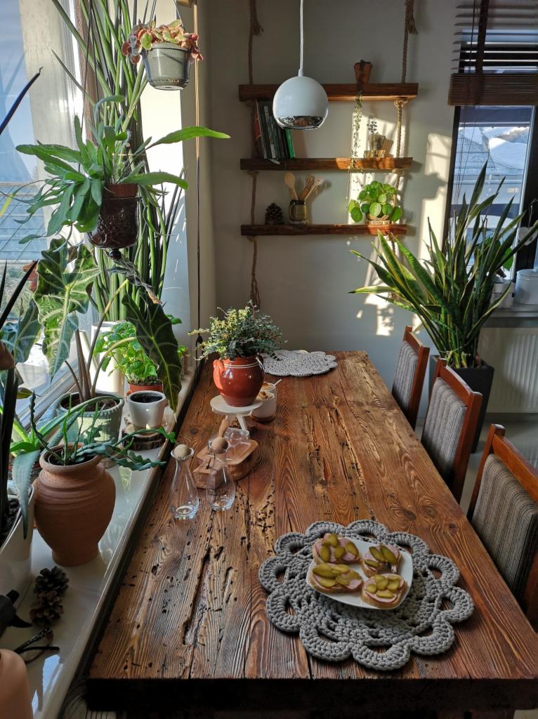 Rustykalny stół z litego drewna, otoczony roślinami doniczkowymi w słonecznym wnętrzu, z widocznymi śladami użytkowania i dekoracyjnymi podkładkami pod talerze.