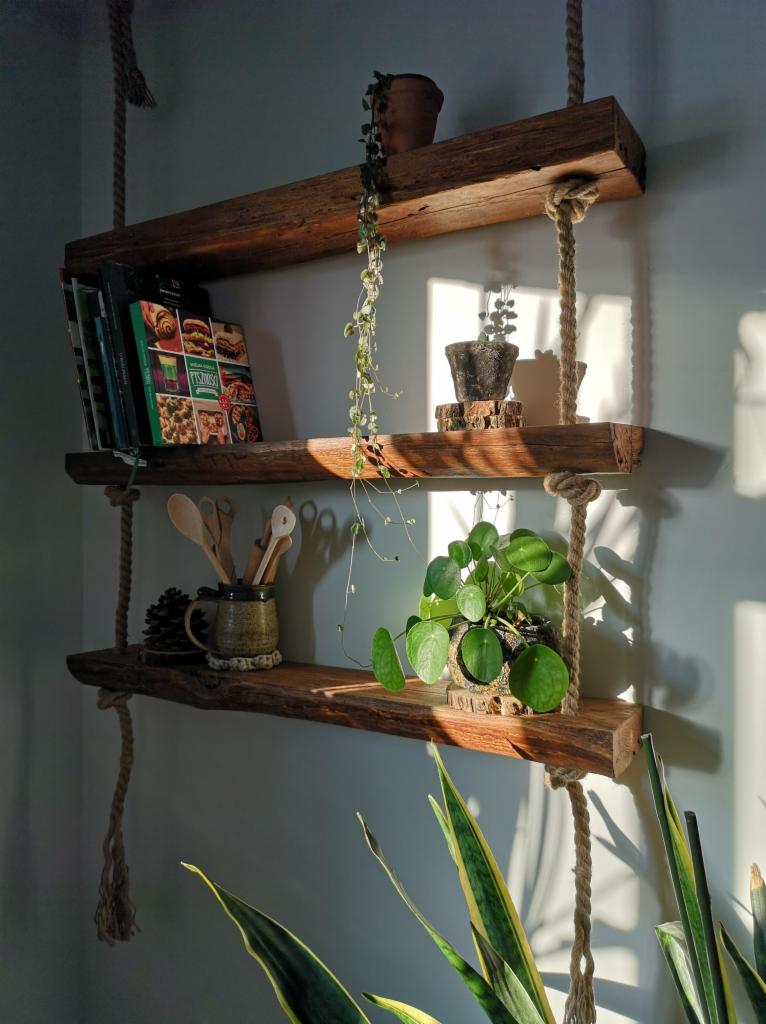 Trzy drewniane półki zawieszone na grubych linach, z książkami kucharskimi, roślinami doniczkowymi i przyborami kuchennymi, oświetlone popołudniowym słońcem tworzącym cienie na ścianie.