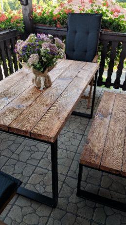Drewniany stół z czarnymi metalowymi nogami i dopasowaną ławką na balkonie, bukiet kwiatów w jutowym worku na stole, widok na zieleń w tle.