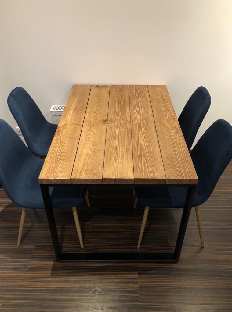 Stół z blatem z jasnego drewna klejonego z desek, na czarnych metalowych nogach, otoczony czterema krzesłami tapicerowanymi granatowym welurem na jasnych drewnianych nogach, na podłodze z paneli...