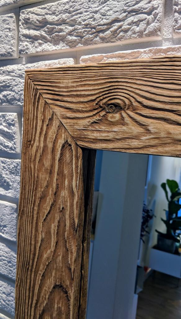 Rama lustra z ciemnego, postarzanego drewna o wyraźnym usłojeniu, na tle białej ściany z cegły dekoracyjnej, z widocznym fragmentem wnętrza z rośliną doniczkową.