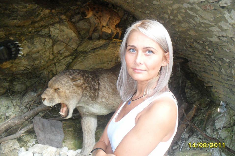 Kobieta w grocie z ekspozycją zwierząt: lew z otwartą paszczą, hiena i niedźwiedzia łapa. Nietypowe tło i oświetlenie.