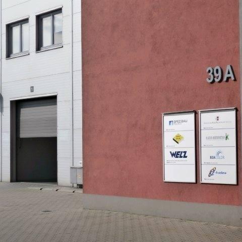 Fasada budynku magazynowego z numerem 39A i tablicami informacyjnymi firm Spec-Bau, Welz, Biacolor i Kalesa.