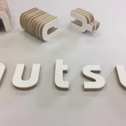 Wycięte laserowo, trójwymiarowe litery z białego tworzywa z widoczną warstwą wewnętrzną z brązowego drewna, tworzące napis 'nutsy', z dodatkowymi literami w tle, częściowo złożonymi.