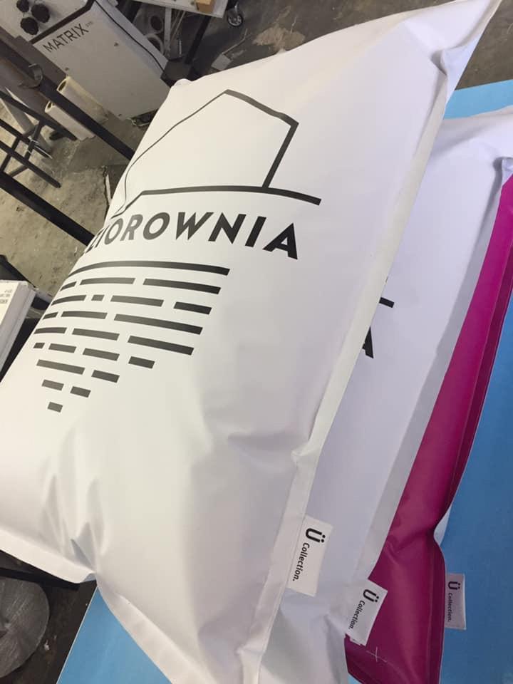 Dwa białe, foliowe opakowania kurierskie z nadrukowanym czarnym logo firmy 'U BOROWNIA' i znakiem 'ü Collection', jedno z różowym akcentem, leżące na niebieskim podłożu, widoczne elementy maszyny...