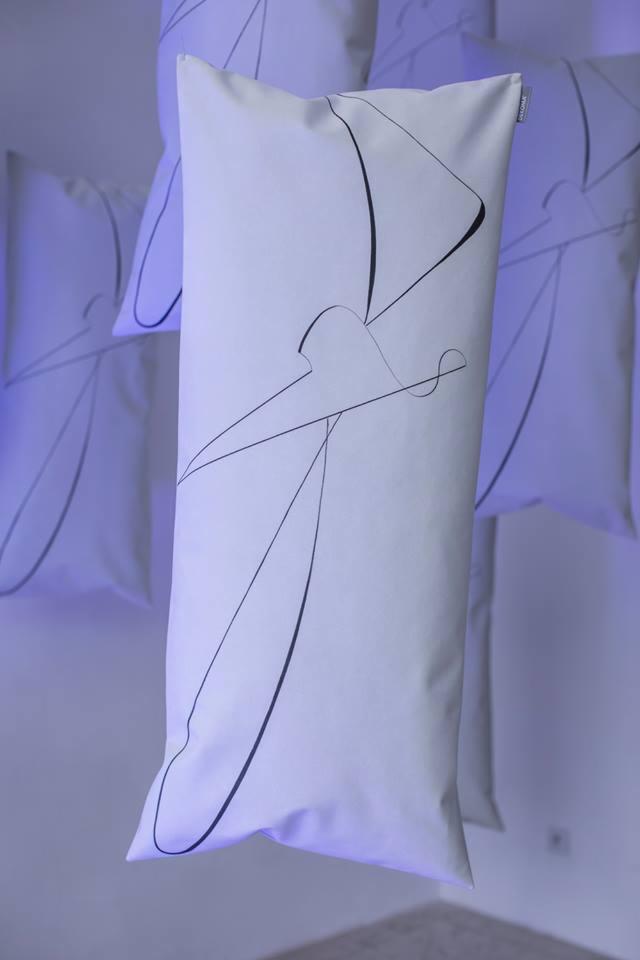 Białe, podłużne poduszki z minimalistycznym czarnym nadrukiem abstrakcyjnym, zwisające w przestrzeni oświetlonej fioletowym światłem.