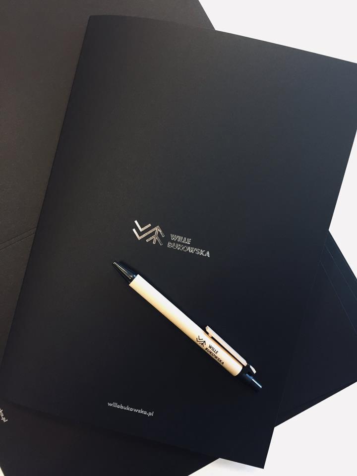 Czarne matowe foldery firmowe z tłoczonym srebrnym logo Wille Bukowska, leżące jeden na drugim, z długopisem reklamowym z tym samym logo na wierzchu.