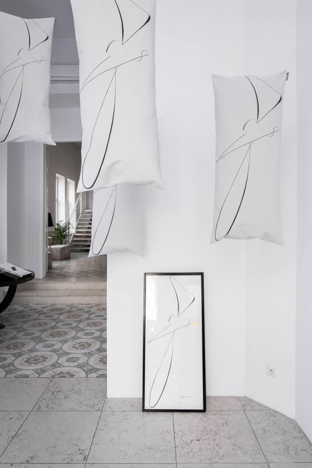 Wnętrze z białymi poduszkami z abstrakcyjnym czarnym nadrukiem wiszącymi pod sufitem i oprawionym plakatem z podobnym wzorem stojącym na podłodze, widok na klatkę schodową w tle.