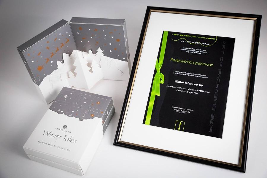 Pudełka na czekoladki z motywem zimowym, otwarte pudełko z wyłaniającymi się choinkami oraz nagroda 'Perła wśród opakowań' w czarnej ramce z zieloną wstążką.