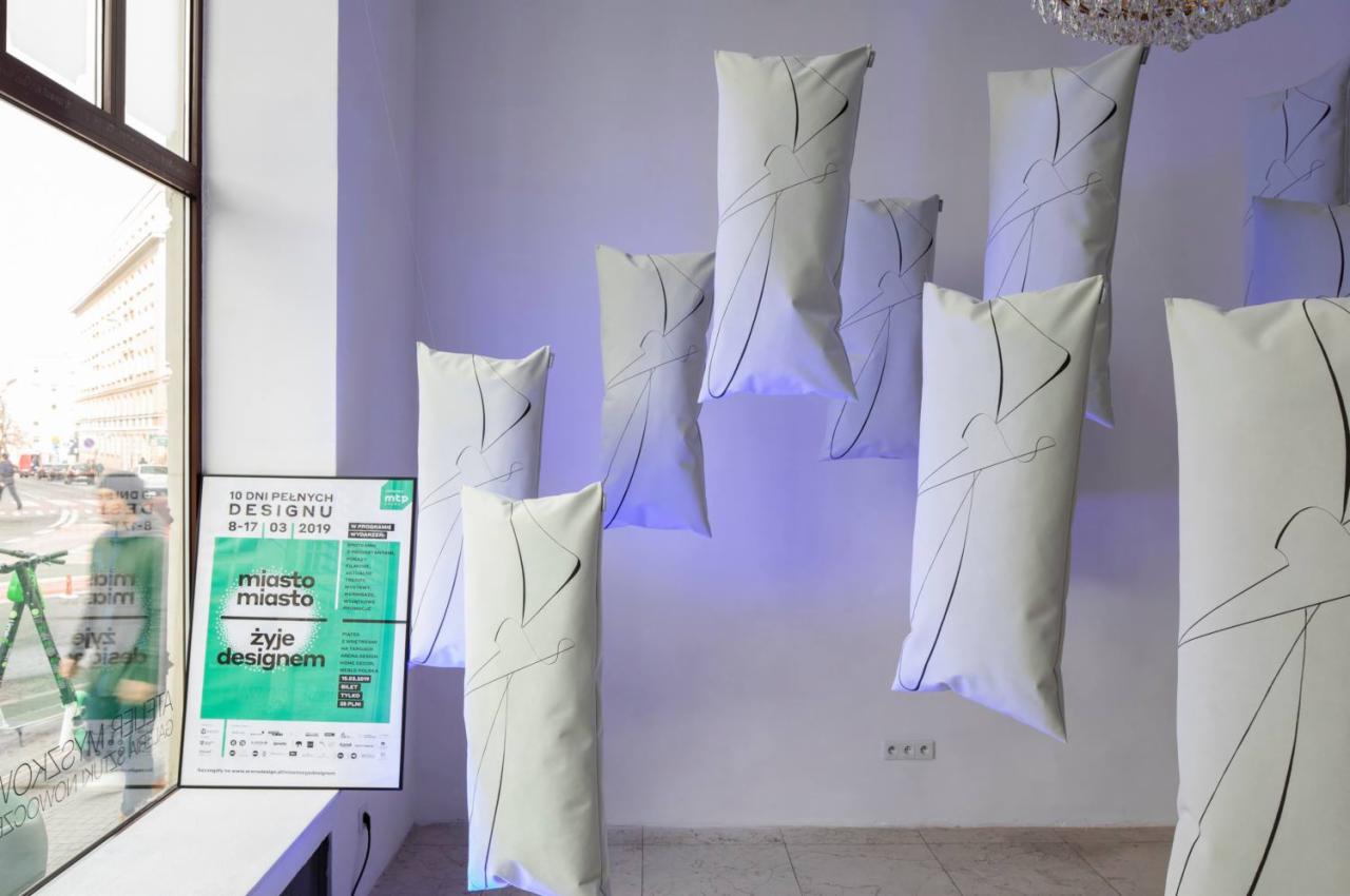 Instalacja artystyczna: białe, podłużne poduszki z czarnym, abstrakcyjnym nadrukiem zawieszone w przestrzeni galerii, plakat informacyjny o wydarzeniu designerskim w tle, widok na ulicę przez okno.