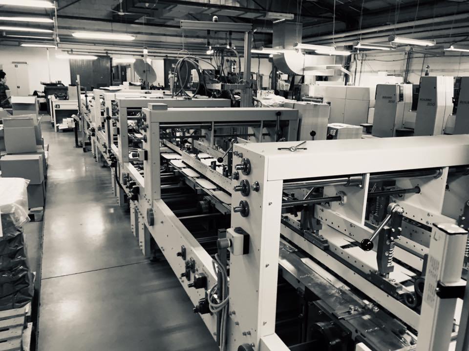 Czarno-białe zdjęcie hali produkcyjnej z długą linią maszyn drukarskich, z widocznymi arkuszami papieru w trakcie obróbki, oświetlenie sufitowe i pracownik w tle po lewej.