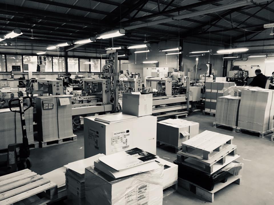 Czarno-białe zdjęcie hali produkcyjnej z maszynami drukarskimi, paletami i kartonami, widoczne osoby obsługujące maszyny.