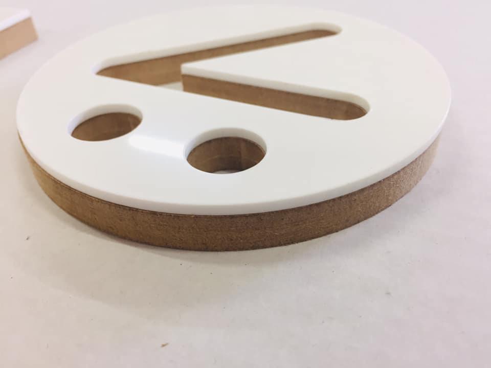 Okrągły, drewniany podkład z wyciętym białym, plastikowym wzorem z dwoma otworami i symbolem 'mniejsze niż'.