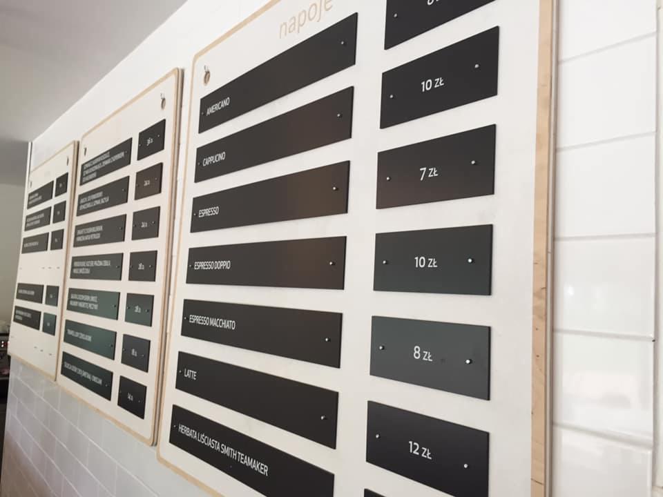 Menu kawiarni wydrukowane na czarnych tabliczkach przytwierdzonych do drewnianej płyty, widoczne nazwy napojów: Americano, Cappuccino, Espresso, Espresso Doppio, Espresso Macchiato, Latte, Herbata...