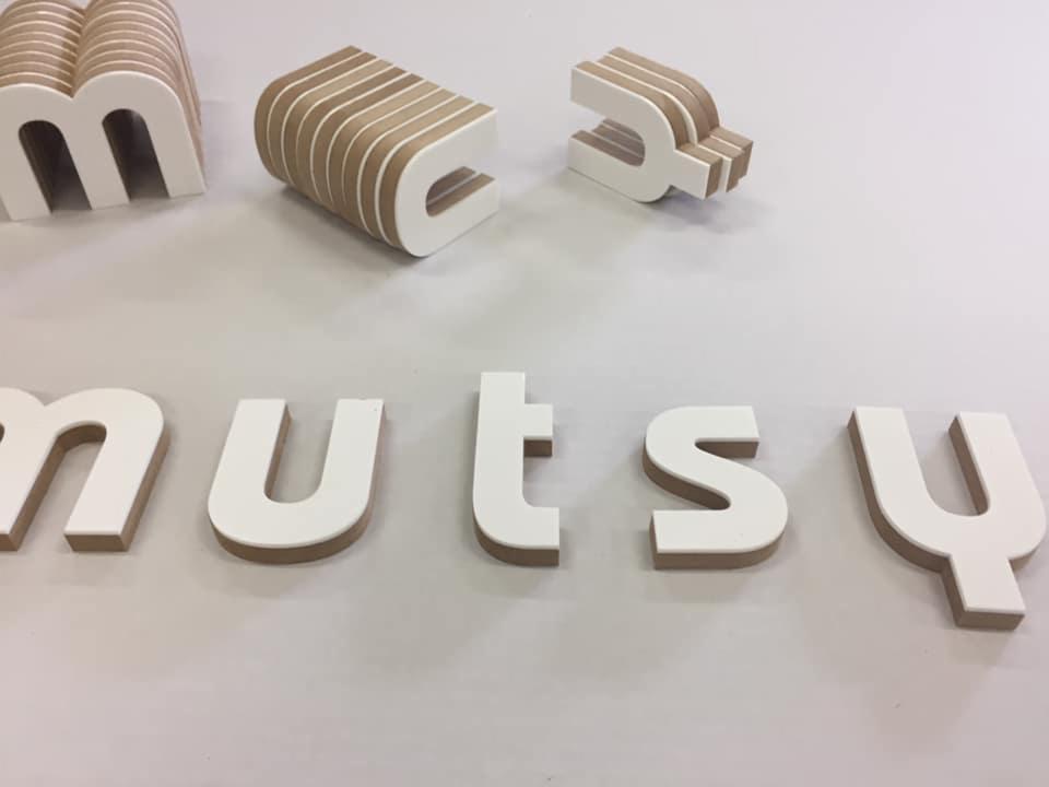Wycięte laserowo, trójwymiarowe litery z białego tworzywa z widoczną warstwą wewnętrzną z brązowego drewna, tworzące napis 'nutsy', z dodatkowymi literami w tle, częściowo złożonymi.