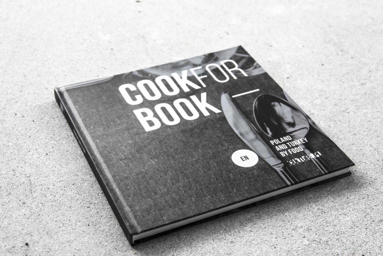 Kwadratowa książka kucharska z napisem COOKFOR BOOK, leżąca na szarym tle z widoczną fakturą, z grafiką sztućców i napisem POLAND AND TURKEY BY FOOD.