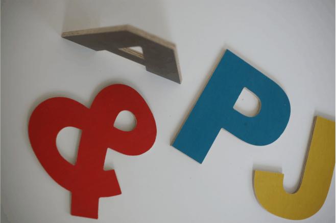 Kolorowe, wycięte litery z drewna (A, B, P, J) na białym tle, ułożone w przypadkowej kolejności, z widoczną fakturą drewna na krawędzi litery A.