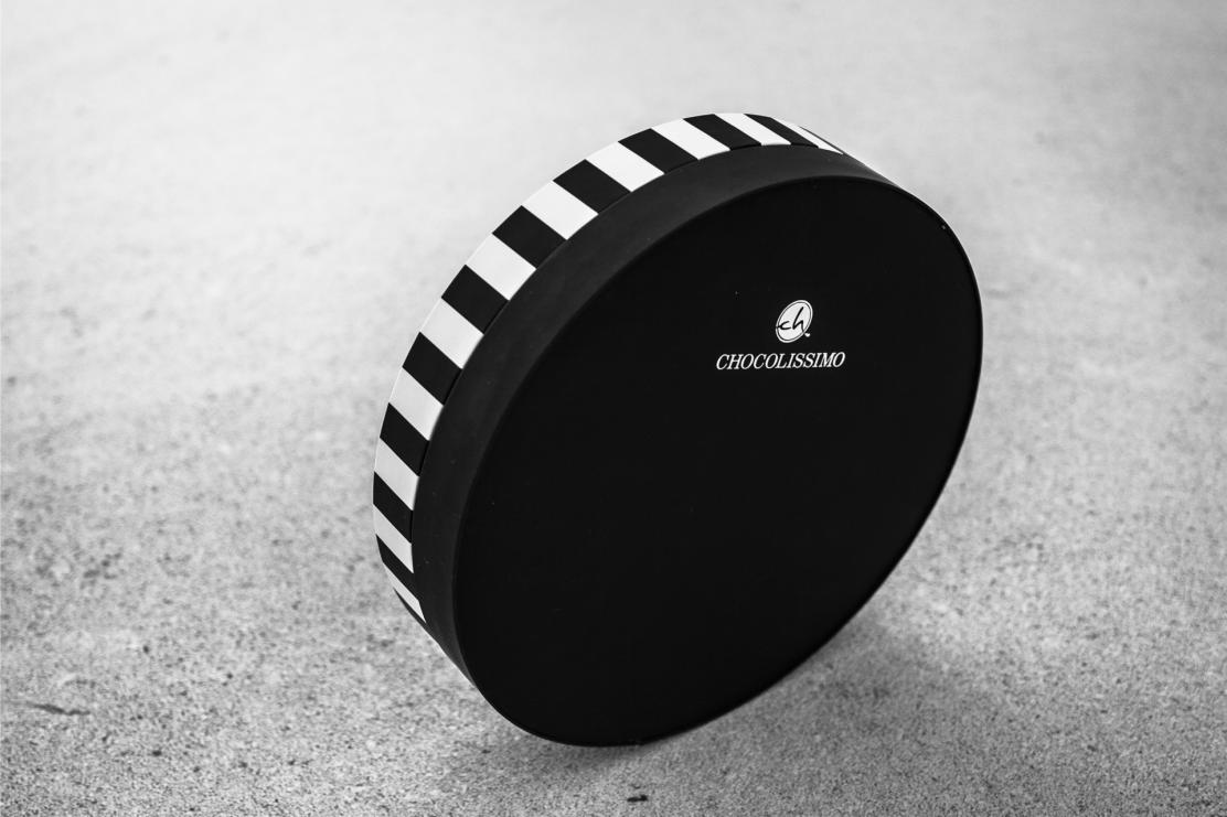 Czarno-białe okrągłe pudełko z paskami na boku i logo 'Chocolissimo', ustawione pod kątem na szarej powierzchni.