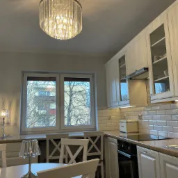 Jasna kuchnia w stylu klasycznym z białymi szafkami, białymi płytkami metra na ścianie, oświetleniem LED pod szafkami i kryształowym żyrandolem, widok na drzewa za oknem