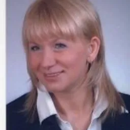 Portret kobiety z blond włosami i niebieskimi oczami, ubrana w białą koszulę i ciemną marynarkę, uśmiechająca się lekko do kamery na tle jasnoszarego tła.