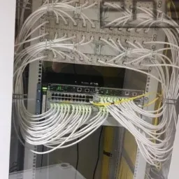 Szafa telekomunikacyjna z patch panelem i switchem, liczne białe kable uporządkowane w wiązki, kilka żółtych kabli, oświetlenie LED na switchu.