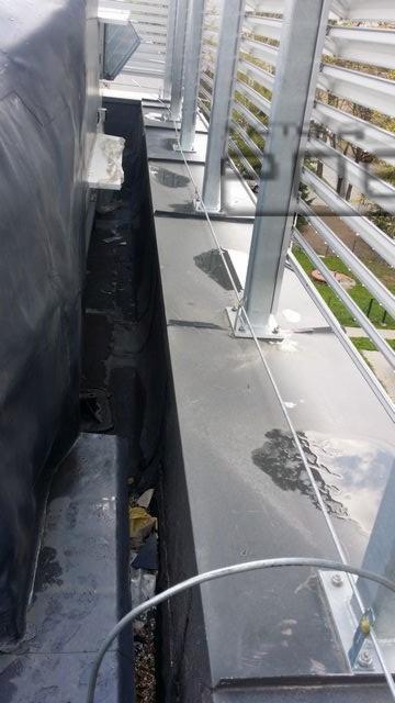 Metalowa konstrukcja wsporcza na dachu z widocznym systemem odwadniania i elementami instalacji odgromowej, skierowana w stronę budynku z żaluzjami.