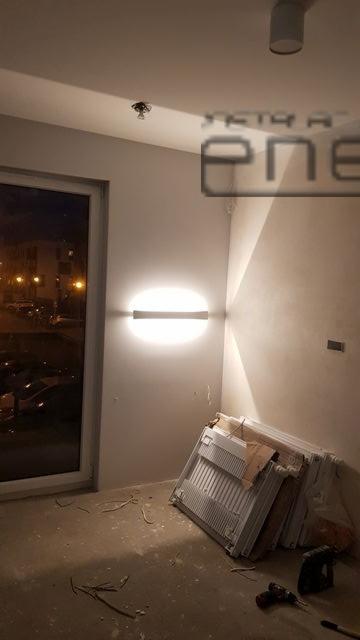 Świecąca, owalna lampa ścienna w surowym wnętrzu, widoczne przygotowane grzejniki i wiertarka na podłodze.