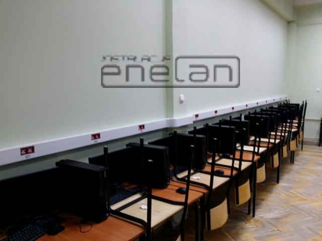 Sala komputerowa z rzędem stanowisk, listwy przypodłogowe z gniazdami elektrycznymi wzdłuż ściany, widok z boku na ustawione w szeregu biurka i krzesła.