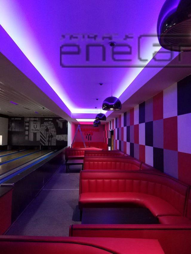 Wnętrze lokalu rozrywkowego z oświetleniem LED w kolorze fioletowym, widoczne czerwone loże, dekoracyjna ściana w szachownicę i tor do gry w kręgle w tle.