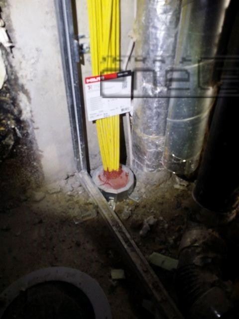 Przejście kablowe przez ścianę, zabezpieczone ogniochronną masą uszczelniającą Hilti, widoczne kable w żółtej izolacji i rury wentylacyjne w tle.