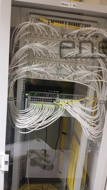 Szafa telekomunikacyjna z patch panelem i switchem, liczne białe kable uporządkowane w wiązki, kilka żółtych kabli, oświetlenie LED na switchu.