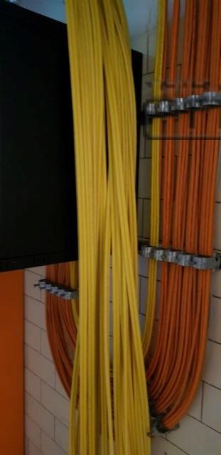 Żółte i pomarańczowe kable ułożone pionowo, przymocowane metalowymi uchwytami do ściany z jasnych, kwadratowych płytek, widoczny fragment czarnej szafki po lewej stronie.