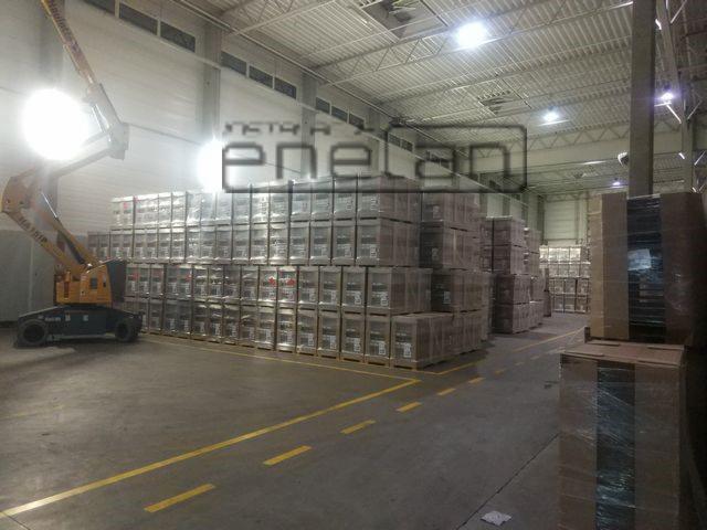 Wnętrze magazynu z paletami opakowanych produktów, żółta podłoga i podnośnik koszowy z oświetleniem w lewym górnym rogu.