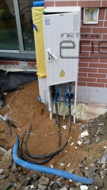 Biała skrzynka elektryczna z otwartymi drzwiczkami, wystającymi kablami w niebieskich peszlach, umieszczona na piaskowym podłożu przy ceglanej ścianie budynku.