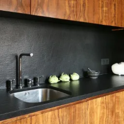 Nowoczesna kuchnia z blatem z ciemnego kamienia o wyraźnej fakturze, zlew ze stali nierdzewnej z minimalistyczną baterią, drewniane szafki z widocznym usłojeniem, trzy zielone warzywa i kamienny...