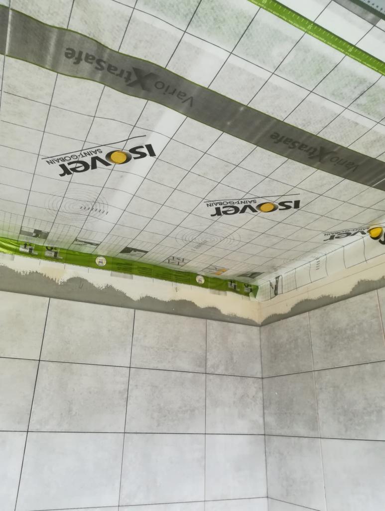 Montaż folii paroizolacyjnej Isover Vario XtraSafe pod płytkami w łazience, widoczne szare płytki na ścianach.