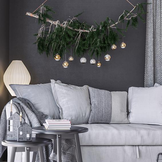 Szaro-biały salon z dekoracją z gałęzi i światełek nad sofą, stolik kawowy z książkami i dekoracyjnym domkiem, lampa podłogowa z lewej strony, zasłona w geometryczny wzór z prawej.